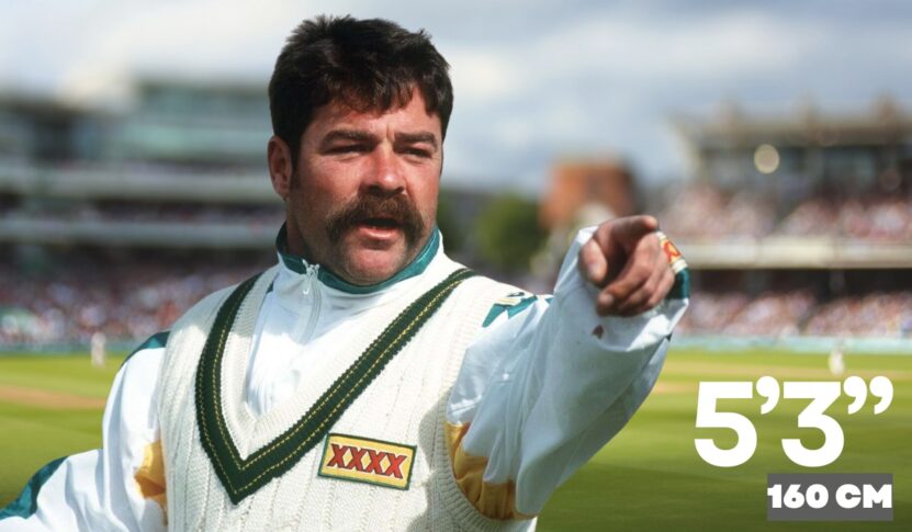 An Australian cricketer in a white sweater with green and gold trim points while standing on a cricket field