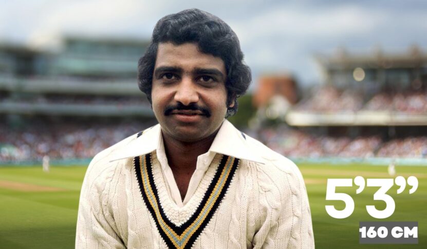 An Indian cricketer wearing a cream-colored sweater with black and yellow trim stands on a cricket field