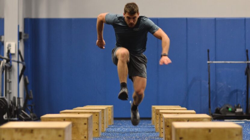 Man performing an agility drill by jumping over wooden boxes in a gym.