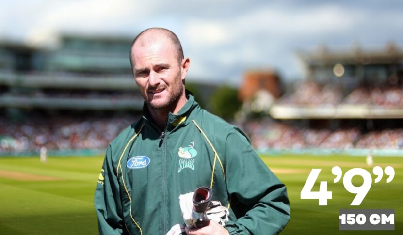 A professional cricketer in a green team jacket holding gloves, standing on a cricket field with a crowd in the background.