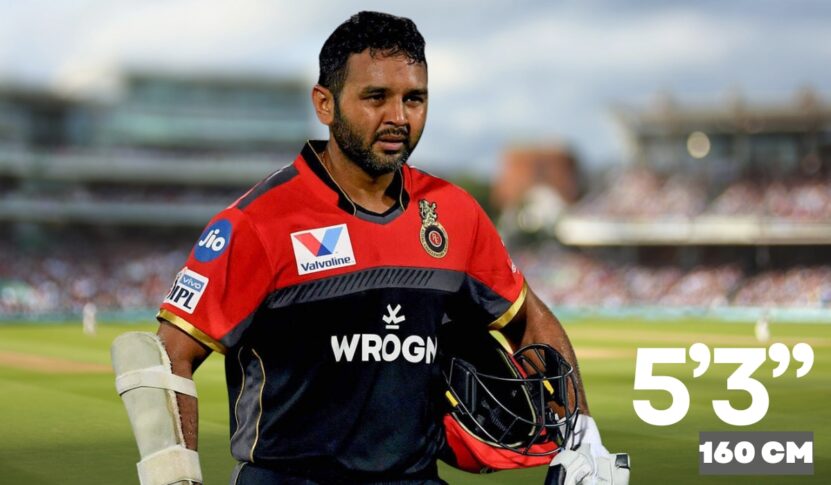 An Indian cricketer in a black and red RCB jersey walks on the cricket field holding his helmet and gloves