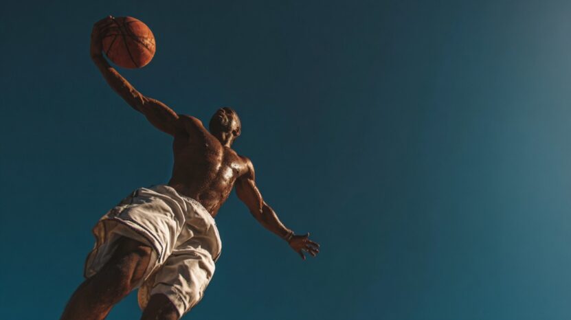 A shirtless athlete in midair holds a basketball overhead against a clear blue sky, captured from a low angle to emphasize height and strength