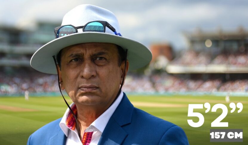 A man in a blue suit, white hat, and sunglasses around his neck stands on a cricket field