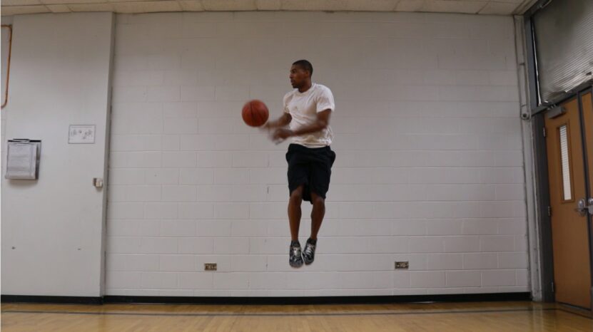 A person in a white shirt and black shorts jumps indoors while holding a basketball, showing strong athletic form and midair control against a white brick wall background