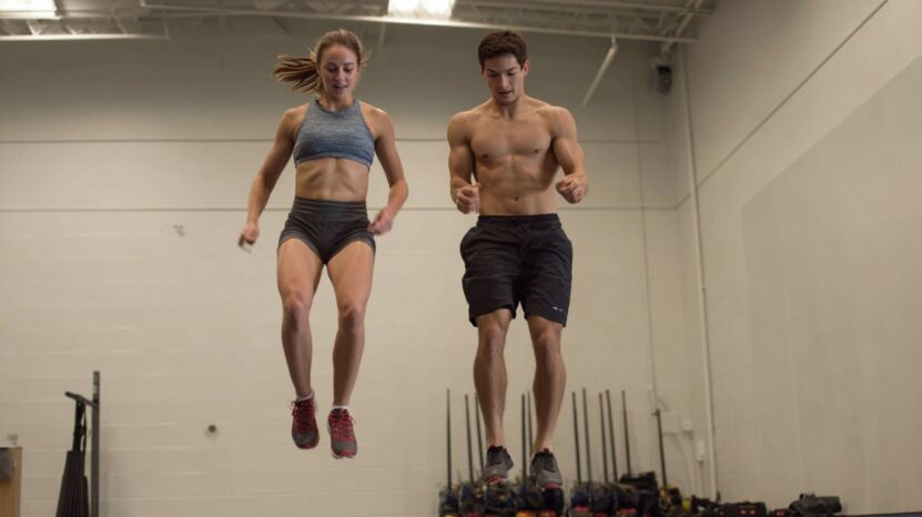 A woman and a man in athletic gear are midair during a jump exercise inside a gym, demonstrating strength and control in their movements