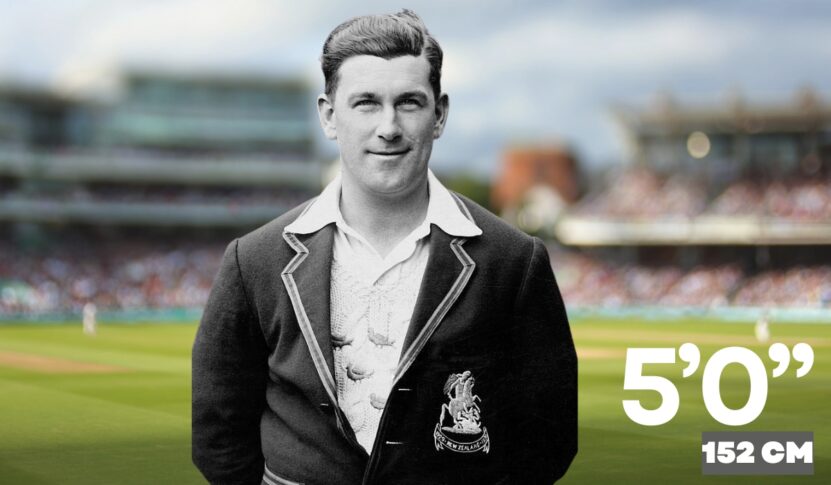 Historic photo of a professional cricketer wearing a dark blazer with a crest, standing on a cricket field with blurred spectators in the background.